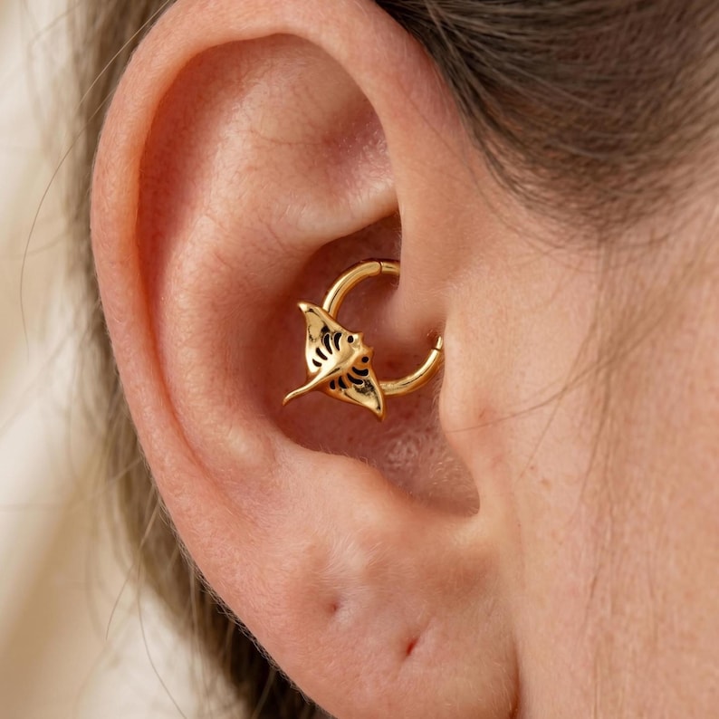May include: A gold-colored daith piercing hoop with a detailed manta ray charm. The charm has intricate cut-out designs and is positioned within the ear's cartilage. The jewelry is shown in a close-up shot.