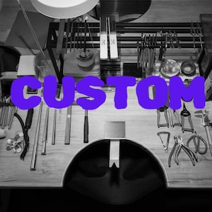 Peut inclure: Un établi avec divers outils de fabrication de bijoux, notamment des pinces, des marteaux et des instruments de mesure. Le mot "CUSTOM" est écrit en grandes lettres violettes sur l'image, suggérant des services de bijoux personnalisés.
