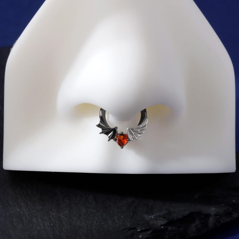 K&ouml;nnte beinhalten: Ein schwarzer Nasenring mit einem herzf&ouml;rmigen roten Edelstein. Der Ring hat auf der einen Seite einen Fledermausfl&uuml;gel und auf der anderen Seite einen wei&szlig;en Fl&uuml;gel. Der Schmuck wird auf einer wei&szlig;en Nasenform pr&auml;sentiert.