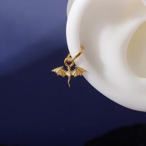 May include: A gold-coloured earring featuring a dragon pendant. The dragon has outstretched wings and a small gemstone. The earring is attached to a gold hoop. The background is a dark blue.