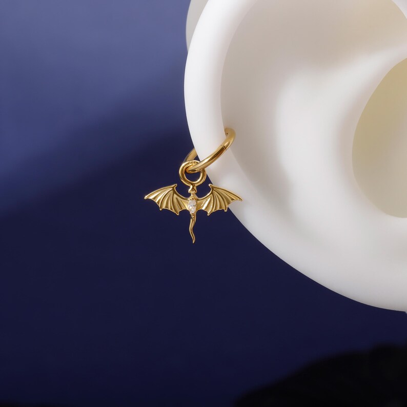 May include: A gold-coloured earring featuring a dragon pendant. The dragon has outstretched wings and a small gemstone. The earring is attached to a gold hoop. The background is a dark blue.