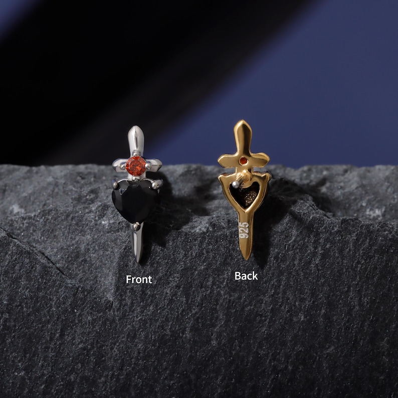 May include: Two small dagger-shaped pendants. One is silver with a black heart-shaped stone and a red gem. The other is gold-colored with a red gem and a heart-shaped cutout. The word "Front" and "Back" are printed on the surface.