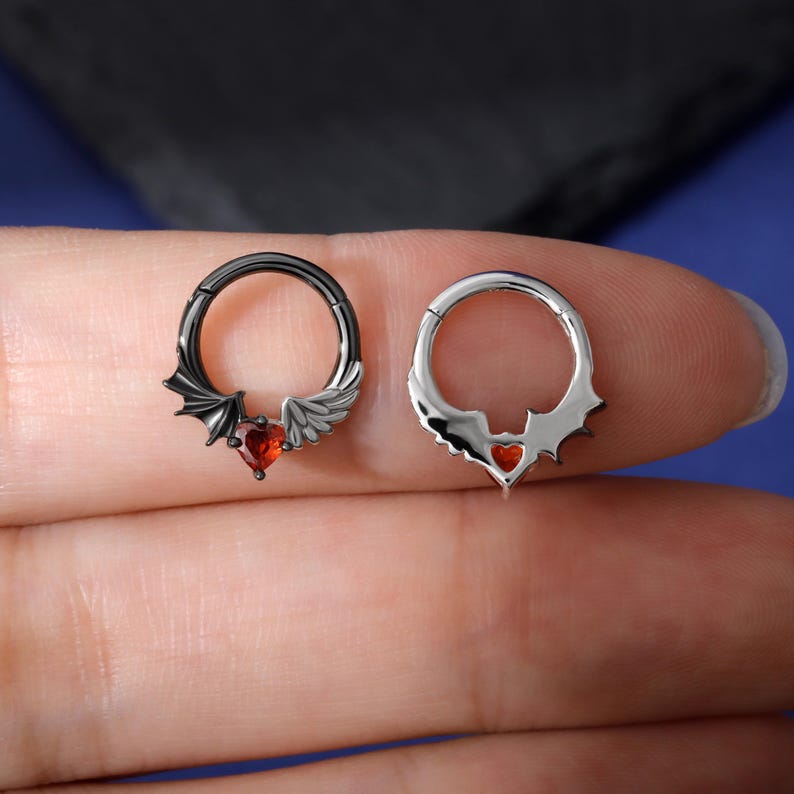 K&ouml;nnte beinhalten: Zwei Septumringe. Einer ist schwarz mit einem Fledermausfl&uuml;gel- und Engelsfl&uuml;gel-Design und einem roten herzf&ouml;rmigen Edelstein. Der andere ist silberfarben mit einem herzf&ouml;rmigen Edelstein und einem dekorativen Rand. Schmuck.