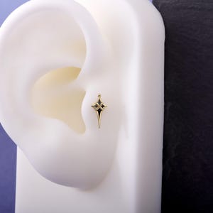 May include: A gold-toned ear stud shaped like a four-pointed star with elongated tips. The centre of the star features a cross-shaped cutout with black enamel detailing, and a small spherical bead sits at the very top point of the star design.