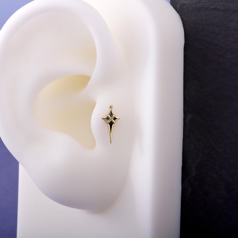 May include: A gold-toned ear stud shaped like a four-pointed star with elongated tips. The centre of the star features a cross-shaped cutout with black enamel detailing, and a small spherical bead sits at the very top point of the star design.
