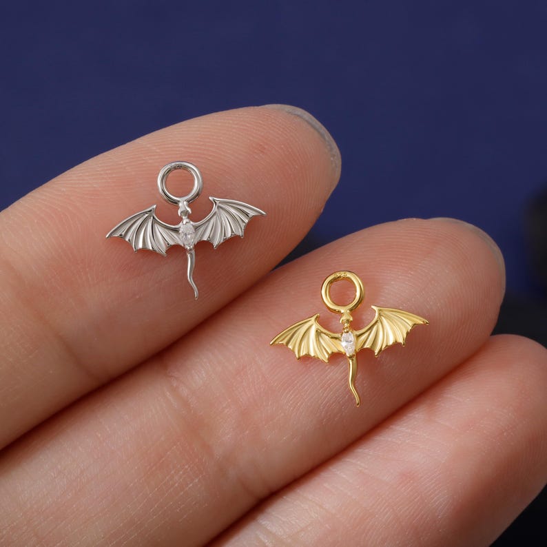 May include: Two small dragon charms, one silver and one gold, each with a small gemstone. The charms feature detailed wings and a loop for attaching to jewelry. The dragon charms are held between fingers against a dark blue background.
