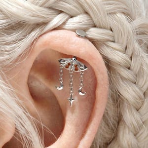May include: A silver-toned moth-shaped cartilage earring featuring two small orange stones on the wings. Three delicate silver chains hang from the moth, with two small crescent moons and a central four-pointed star charm dangling at the ends.