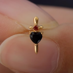 May include: A gold-colored earring with a dagger design. The earring features a black heart-shaped stone at the bottom, a red round stone in the middle, and a cross-shaped top. The earring is designed to be worn in a piercing.