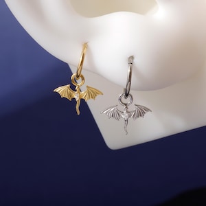 Könnte beinhalten: Zwei Creolen, eine goldfarbene und eine silberfarbene, jeweils mit einem Drachenanhänger. Die Drachenanhänger haben ausgebreitete Flügel und einen kleinen, klaren Edelstein in der Mitte. Die Ohrringe werden vor einem weißen Hintergrund mit dunkelblauem Hintergrund gezeigt.