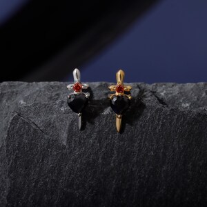 May include: Two dagger-shaped earrings. One is silver-toned with a black heart-shaped gem and a red gem at the top. The other is gold-toned with a black heart-shaped gem and a red gem at the top. The earrings are displayed on a dark gray, textured surface.