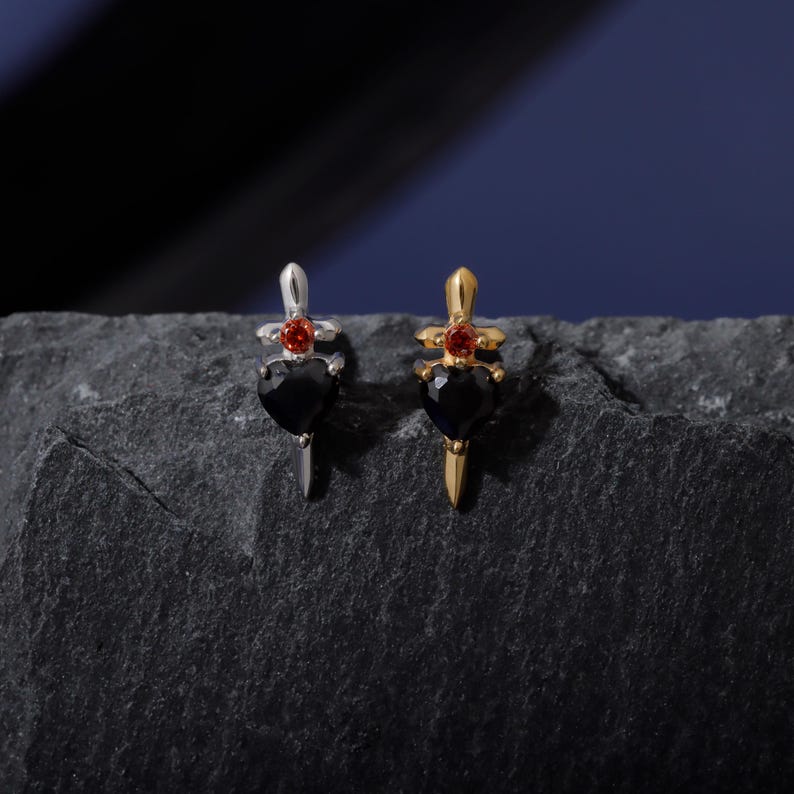 May include: Two dagger-shaped earrings. One is silver-toned with a black heart-shaped gem and a red gem at the top. The other is gold-toned with a black heart-shaped gem and a red gem at the top. The earrings are displayed on a dark gray, textured surface.