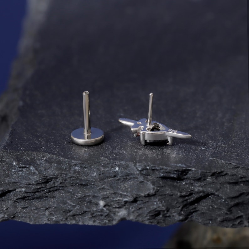 May include: A pair of silver-colored stud earrings. One earring is a post with a round backing, and the other is a stylized bee shape with a black stone center. The earrings are displayed on a dark, textured surface.