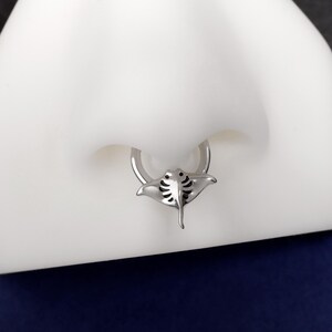 May include: A silver-colored septum ring featuring a manta ray design. The ring is attached to a white surface, with a dark blue background. The manta ray has detailed wing and body features.