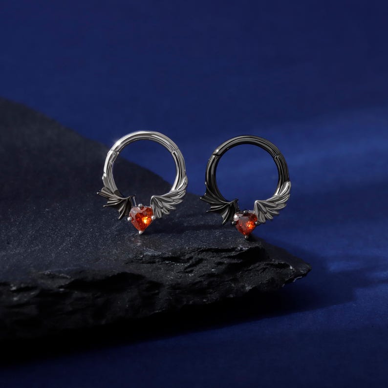 K&ouml;nnte beinhalten: Zwei silberne und schwarze Creolen mit herzf&ouml;rmigem rotem Edelstein und Fl&uuml;geldesign. Die Ohrringe werden auf einer dunklen, strukturierten Oberfl&auml;che vor einem tiefblauen Hintergrund pr&auml;sentiert. K&ouml;rperschmuck.