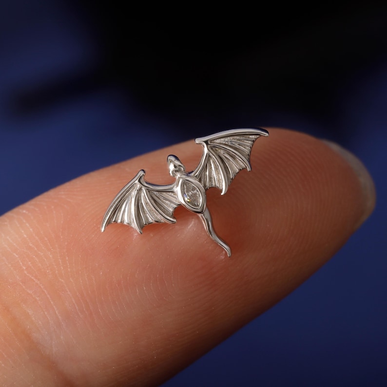 May include: A small, silver-colored dragon pendant with outstretched wings and a gemstone in the center of its body. The dragon is detailed with textured wings and a slender tail. The pendant is shown on a fingertip against a dark blue background.