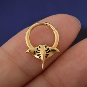 May include: A gold-colored hinged ring with a manta ray design. The manta ray has a detailed pattern on its wings and a pointed tail. The ring is likely for body piercing.