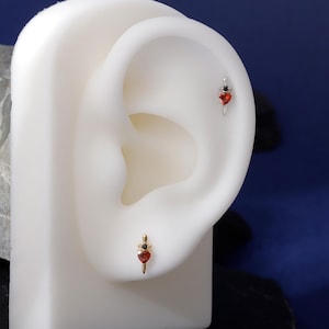 Puede incluir: Dos pendientes exhibidos en un modelo de oreja blanco. Un pendiente es dorado con una piedra roja en forma de corazón y una piedra negra. El otro pendiente es plateado con una piedra roja en forma de corazón y una piedra negra.