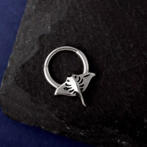 May include: A silver-colored, hinged hoop earring with a manta ray design. The earring features a circular hoop with a detailed manta ray charm attached. The manta ray has a stylized, open-wing design with intricate cutouts. The earring is displayed on a dark, textured surface.