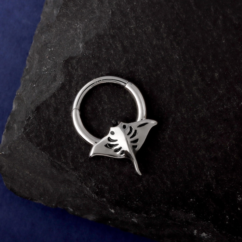 May include: A silver-colored, hinged hoop earring with a manta ray design. The earring features a circular hoop with a detailed manta ray charm attached. The manta ray has a stylized, open-wing design with intricate cutouts. The earring is displayed on a dark, textured surface.