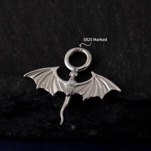 May include: A silver dragon pendant with detailed wings and a loop for a chain. The pendant has a curved tail and etched wing details. The text "S925 Marked" is visible. The pendant is likely made of sterling silver.