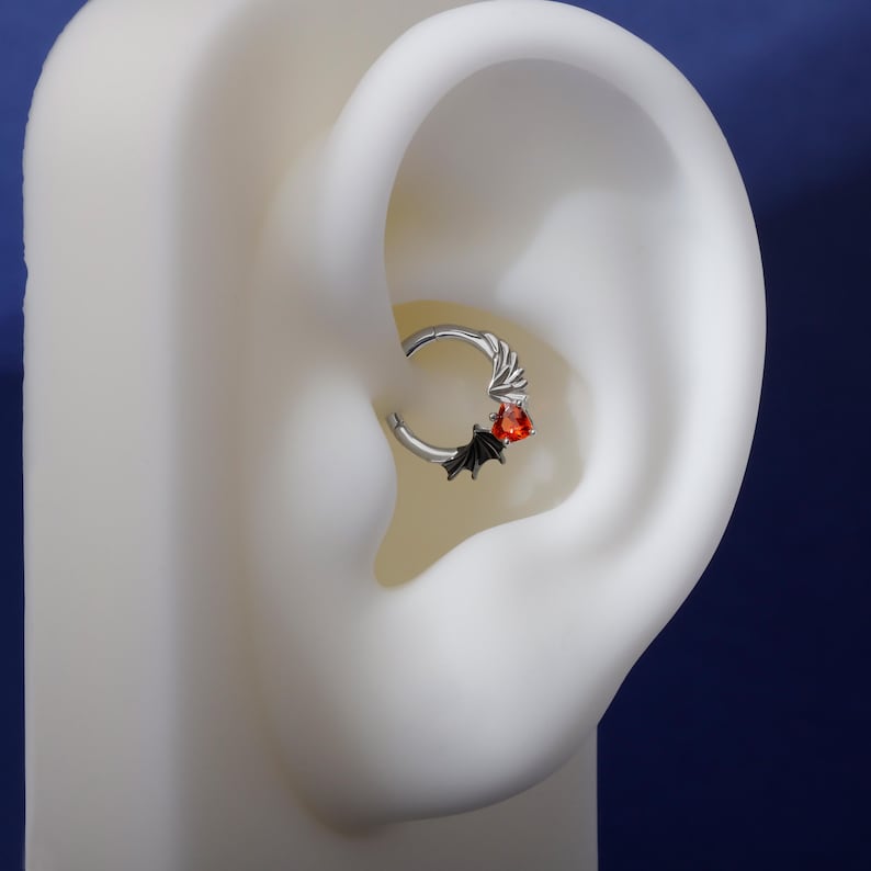 K&ouml;nnte beinhalten: Ein silberner Septumring mit einem Fledermaus- und Edelstein-Design. Der Ring hat eine kleine, schwarze Fledermaus und einen roten Edelstein. Das Design ist vor einem wei&szlig;en Ohrmodell und einem dunkelblauen Hintergrund platziert.