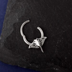 May include: A silver-colored hinged hoop earring featuring a manta ray design. The earring has a detailed cutout of the manta ray's body and wings. The earring is shown against a dark, textured background.