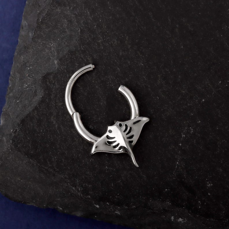 May include: A silver-colored hinged hoop earring featuring a manta ray design. The earring has a detailed cutout of the manta ray's body and wings. The earring is shown against a dark, textured background.