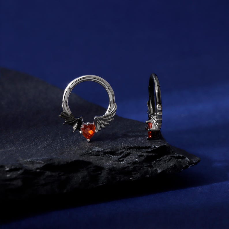 K&ouml;nnte beinhalten: Zwei silberfarbene Creolen. Ein Ohrring hat einen roten herzf&ouml;rmigen Edelstein mit Fledermaus- und Engelsfl&uuml;geln. Der andere Ohrring hat einen kleinen roten Edelstein. Die Ohrringe sind auf dunkler, strukturierter Oberfl&auml;che vor dunkelblauem Hintergrund.