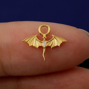 May include: A gold dragon pendant with detailed wings and a central clear gemstone. The dragon has a curved tail and a ring at the top. The pendant is held between two fingers, showing the intricate design.