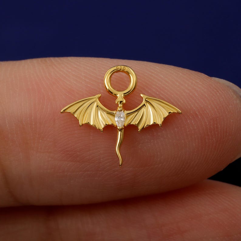 May include: A gold dragon pendant with detailed wings and a central clear gemstone. The dragon has a curved tail and a ring at the top. The pendant is held between two fingers, showing the intricate design.