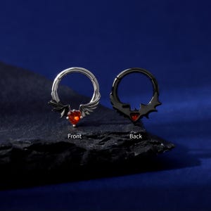 K&ouml;nnte beinhalten: Zwei Septumringe, einer silberfarben und einer schwarz, jeweils mit einem herzf&ouml;rmigen roten Edelstein. Der silberne Ring hat fl&uuml;gelartige Akzente, w&auml;hrend der schwarze Ring Fledermausfl&uuml;gel-Designs hat. Die W&ouml;rter "Front" und "Back" sind beschriftet.