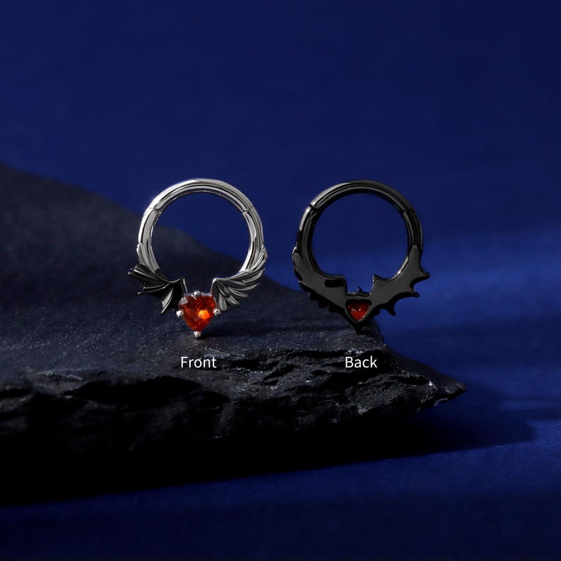 K&ouml;nnte beinhalten: Zwei Septumringe, einer silberfarben und einer schwarz, jeweils mit einem herzf&ouml;rmigen roten Edelstein. Der silberne Ring hat fl&uuml;gelartige Akzente, w&auml;hrend der schwarze Ring Fledermausfl&uuml;gel-Designs hat. Die W&ouml;rter "Front" und "Back" sind beschriftet.