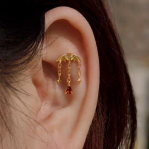 May include: A gold-colored ear piercing featuring a bat design with dangling chains. The chains hold star-shaped charms and a teardrop-shaped red gemstone. The piercing is attached to the ear.