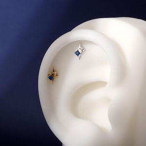 Puede incluir: Primer plano de dos pendientes en forma de estrella con gemas cuadradas azules y pequeñas piedras transparentes. Un pendiente es dorado, el otro es plateado. Los pendientes se muestran en un modelo de oreja blanca sobre un fondo azul oscuro.