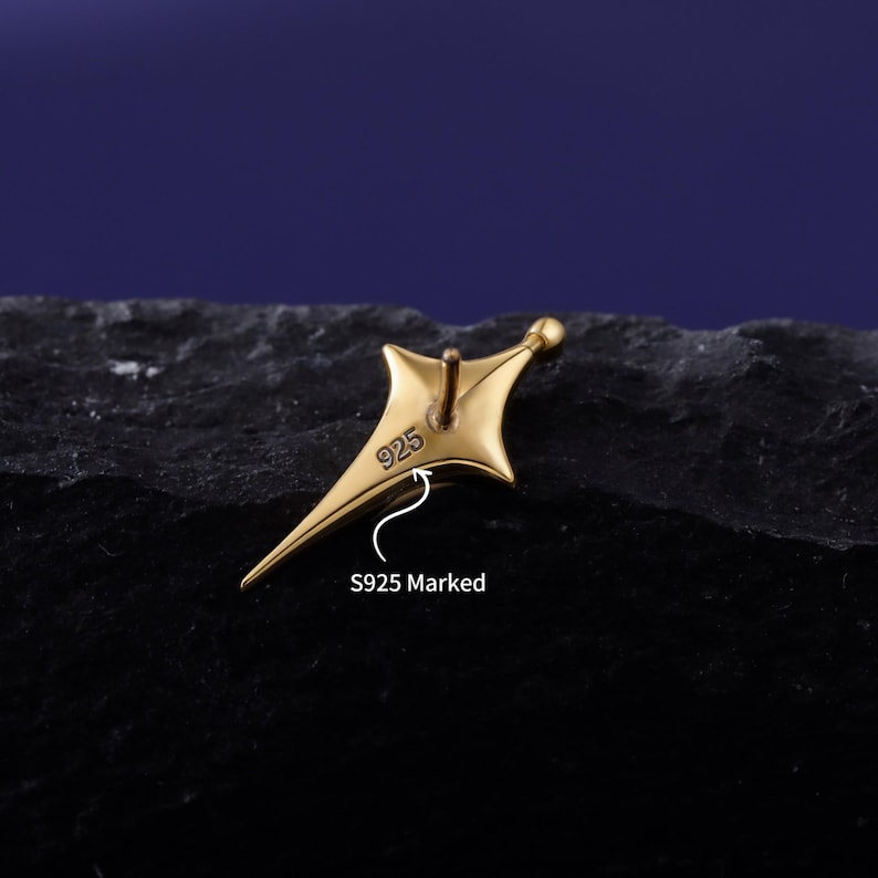 May include: A gold-toned star-shaped earring stud with a pointed design. The back of the earring features a clearly visible 925 stamp, indicating sterling silver composition. A small post extends from the centre of the star-shaped metal piece.