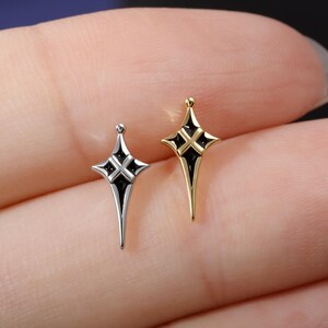 May include: Two small, elongated star-shaped charms, one in a silver-tone metal and one in a gold-tone metal. Each charm features a central cross-shaped cutout filled with black enamel, with a small spherical bead at the top point of the star.