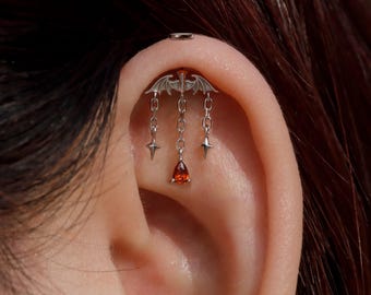 May include: A silver-toned ear cartilage piercing featuring a bat-shaped top piece with three dangling chains. The two outer chains end in four-pointed star charms, while the centre chain features a single teardrop-shaped orange gemstone.