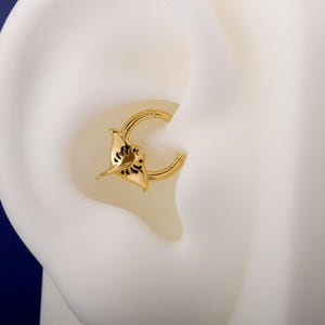 Puede incluir: Pendiente dorado con diseño de manta raya. El pendiente es de tipo aro, con el dije de manta raya adjunto en la parte delantera. La manta raya tiene detalles en negro. El pendiente se muestra sobre un fondo blanco.