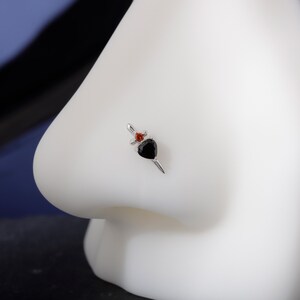 May include: A silver nose stud featuring a black heart-shaped gem and a small red gem. The stud has a dagger design, with the heart as the blade and the red gem at the hilt. The jewelry is set against a white background.
