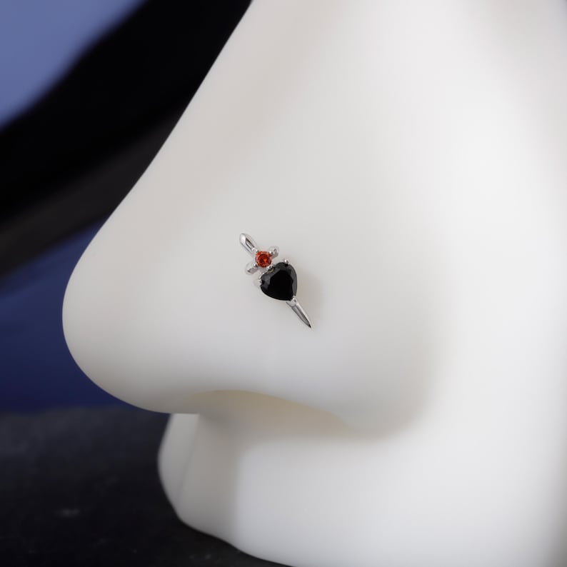 May include: A silver nose stud featuring a black heart-shaped gem and a small red gem. The stud has a dagger design, with the heart as the blade and the red gem at the hilt. The jewelry is set against a white background.