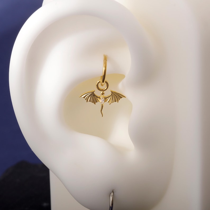 May include: A gold-coloured earring with a dragon pendant. The pendant has outstretched wings and a small gemstone. The earring is a hoop style, designed for a pierced ear. The dragon pendant has detailed wing features.