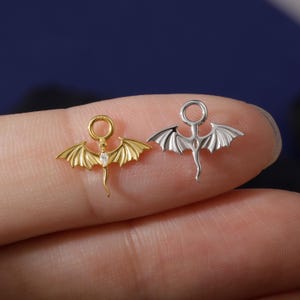 May include: Two small dragon-shaped charms. One is gold-coloured with a clear gem and the other is silver-coloured. Each charm has a loop at the top for hanging and detailed wings. The charms are displayed against a blurred background.