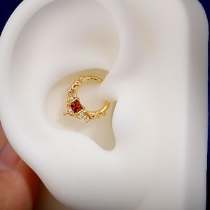 May include: A gold-colored septum ring with a red square gemstone and small white gemstones. The ring has a decorative, curved design. The jewelry is shown in a close-up view, set in a white ear.
