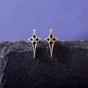 May include: Two star-shaped charms, one in silver-tone metal and one in gold-tone metal, each featuring a pointed, elongated design with black enamel inlay details. The charms are positioned side-by-side, showcasing their symmetrical, four-pointed star shape.