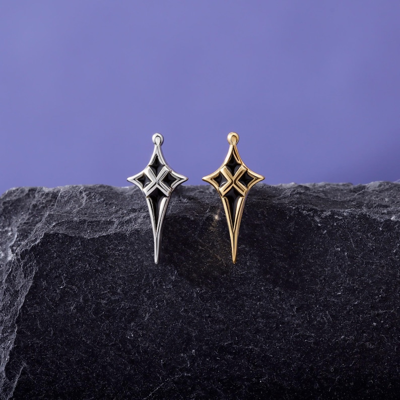 May include: Two star-shaped charms, one in silver-tone metal and one in gold-tone metal, each featuring a pointed, elongated design with black enamel inlay details. The charms are positioned side-by-side, showcasing their symmetrical, four-pointed star shape.