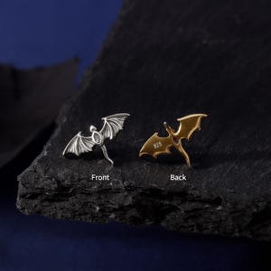 May include: Two dragon-shaped earrings, one silver and one gold, displayed against a dark background. The silver earring shows the front, with intricate wing details. The gold earring, marked "925" on the back, is also visible. The earrings are small and detailed.