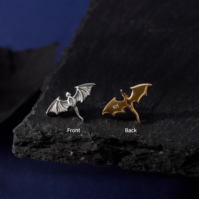 May include: Two dragon-shaped earrings, one silver and one gold, displayed against a dark background. The silver earring shows the front, with intricate wing details. The gold earring, marked "925" on the back, is also visible. The earrings are small and detailed.