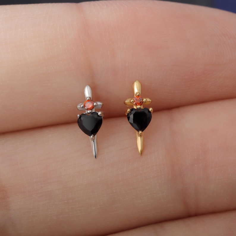 May include: Two small dagger earrings. One is silver-toned with a black heart-shaped gem and a red gem at the top. The other is gold-toned with a black heart-shaped gem and a red gem at the top. The earrings are small and delicate.