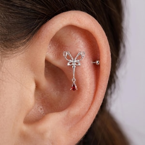May include: A silver-colored ear piercing featuring a bow-shaped top design with a dangling teardrop-shaped, deep red gemstone. The jewelry is shown on an ear, highlighting the intricate details and the gemstone's vibrant color.