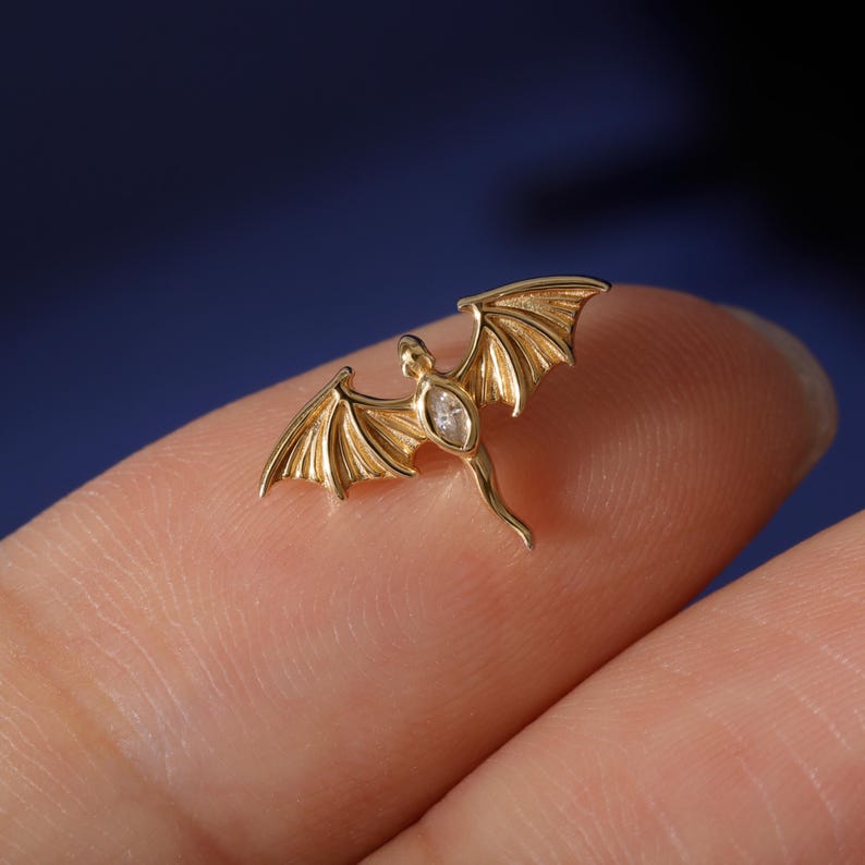 May include: A gold-colored dragon pendant with outstretched wings and a small, clear gemstone in the center. The dragon is detailed with textured wings and a slender body. The pendant is shown against a blurred background.